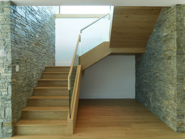 Interior view of a contemporary stairway with warm timber flooring, timber stair treads and angular timber balustrade. Rough stacked-stone walls frame both sides, while a large translucent glazed opening and skylight-like overhead opening bring in natural light. The stair continues beneath a timber-clad structural or landing element, creating a sculptural, highly integrated composition.