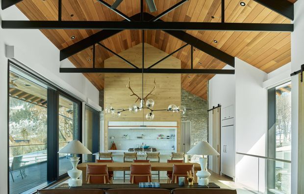 Bright open-plan interior featuring a dining area and living space beneath a high vaulted timber ceiling with exposed dark structural beams. Floor-to-ceiling glazing opens to an outdoor deck, while a stone feature wall, built-in shelving, contemporary pendant lighting, and warm timber finishes define the room.