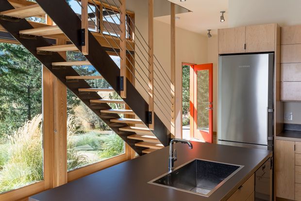 Bright open-plan kitchen interior with a prominent exposed timber-and-steel open-riser staircase, large timber-framed windows, warm light-wood cabinetry, dark benchtops, and a stainless-steel refrigerator. The kitchen sink and island dominate the foreground, while a landscaped garden is visible through the glazing.