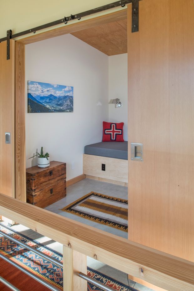A compact, warm-toned sleeping nook or small bedroom viewed through an open sliding barn door. The space features a built-in window-side bench with a grey cushion, light timber joinery and ceiling lining, white walls, a rustic chest of drawers, patterned geometric rugs, a mountain landscape artwork, and a red decorative cushion. Black metal barn-door hardware and a timber-framed glazed balustrade are prominent in the foreground.