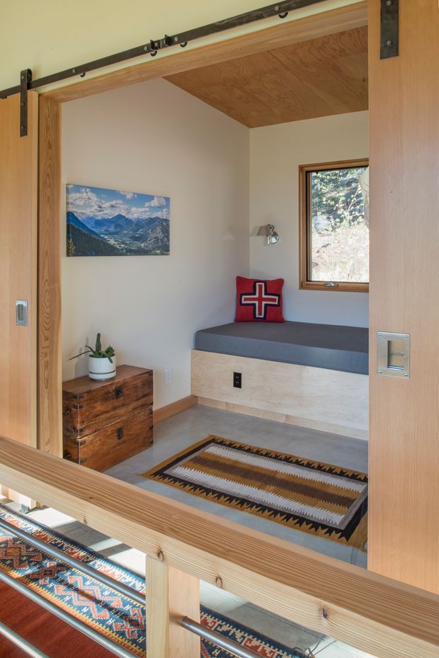 Compact, warmly finished sleeping nook or bedroom viewed through a timber-framed sliding barn door. The room features a built-in window seat/daybed with grey cushion, pale timber base and ceiling lining, white walls, a small timber chest, wall light, framed mountain artwork, and patterned woven rugs. Timber-framed glazing and a timber balustrade are visible in the foreground, giving the space a cabin-like, natural character.