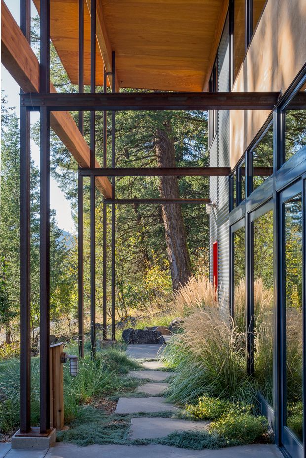 A covered exterior walkway beside a modern building, framed by dark steel posts and beams with a warm timber soffit overhead. Full-height glazing and corrugated metal wall cladding line the right side, reflecting the surrounding wooded landscape. A stepping-stone concrete path passes through ornamental grasses and native-style planting toward mature trees and a mountainous woodland setting.