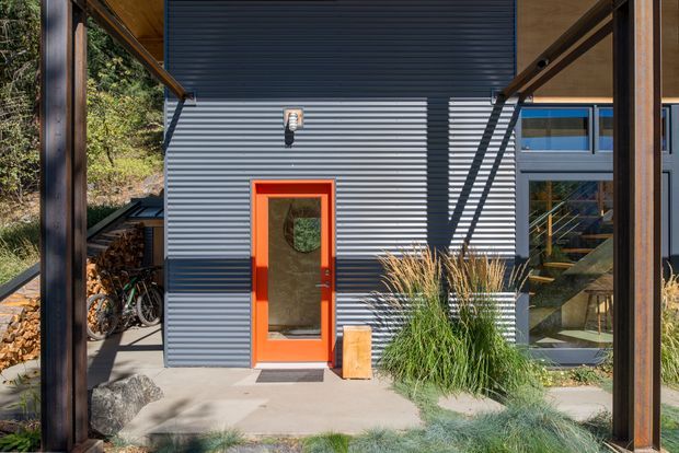 Exterior view of a contemporary compact building framed by dark timber posts and beams. The facade is clad in horizontal dark grey ribbed metal, with a vivid orange entry door, adjacent glazing, and a visible exterior stair. Timber-lined soffits, planted grasses, stone landscaping, bicycles, and stacked firewood contribute to a rustic natural setting.