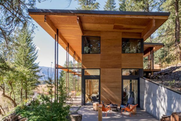 Exterior view of a modern timber-clad house set among trees on a sloping site. The building has broad projecting flat roof planes, extensive dark-framed glazing, a covered terrace with outdoor chairs, and a concrete retaining wall. Mountain terrain is visible in the distance.