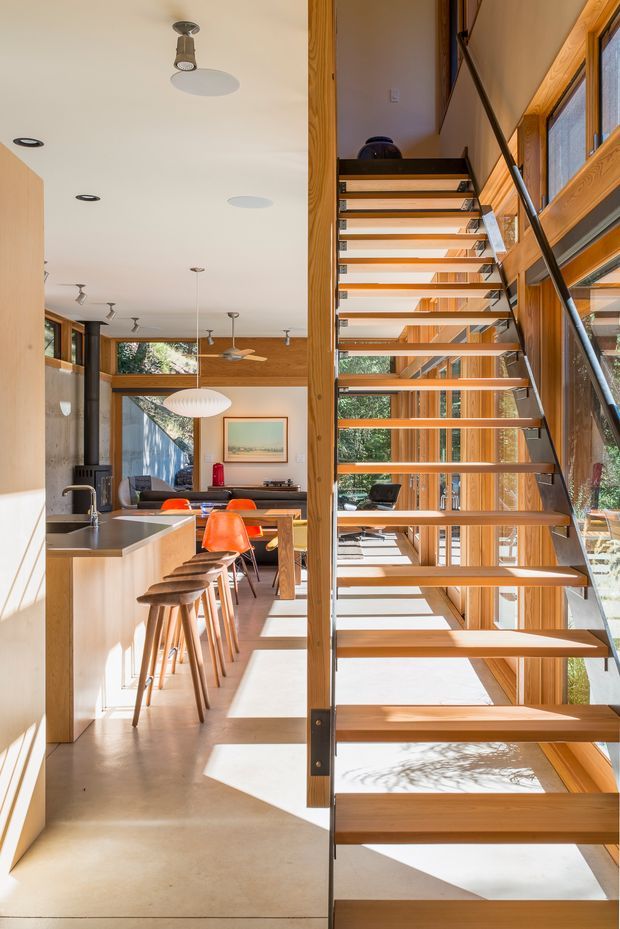 A bright, open-plan contemporary interior viewed from beside a floating staircase. The staircase has open timber treads, slim dark metal stringers, and a dark handrail, with extensive timber-framed glazing along the right side. To the left is a kitchen with a pale timber island, dark benchtop, and timber stools, opening to a dining and living area with colourful dining chairs, pendant lighting, timber wall and ceiling details, and abundant views through the windows. Strong sunlight creates a warm, airy atmosphere.