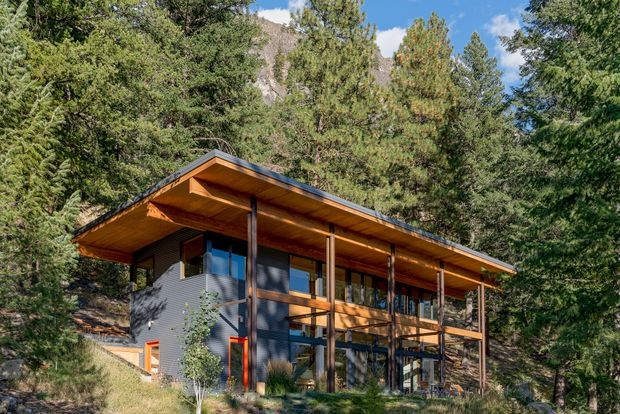 A contemporary two-storey mountain home set among dense forest, with a broad low-pitched projecting roof, warm timber soffits and balcony structure, dark horizontal exterior cladding, and extensive glazing. The elevated deck and slender timber posts face the wooded landscape, with rugged mountains visible behind the trees.