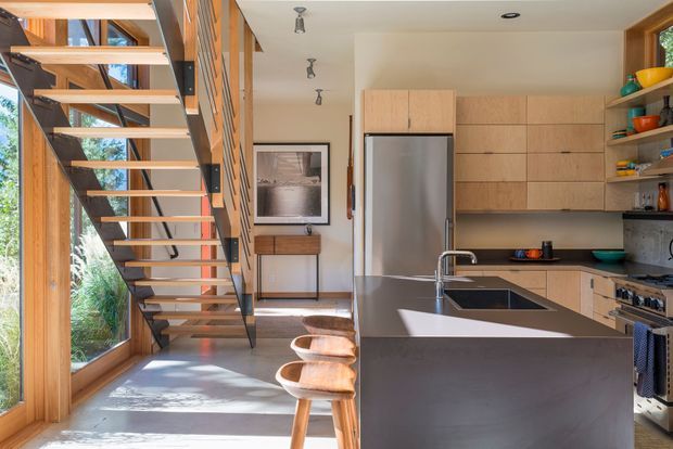 A bright contemporary kitchen viewed beside an open-riser staircase. The kitchen features a large dark island with integrated sink and dishwasher, pale timber cabinetry, open shelving, stainless-steel appliances, and a timber-framed glazed wall bringing in abundant natural light. The staircase combines timber treads with dark metal supports and balustrade elements.