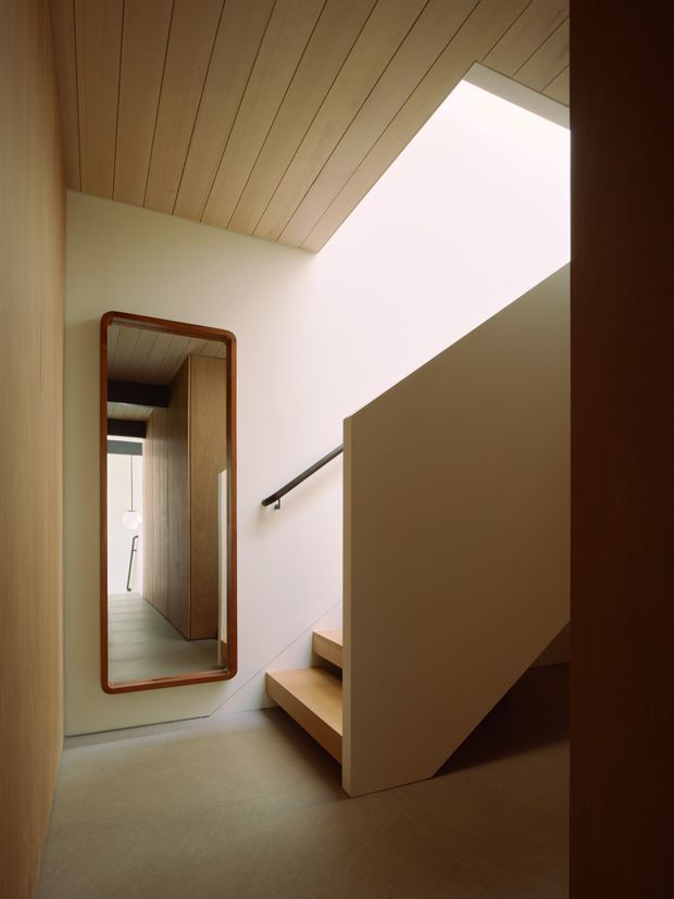 Interior view of a compact, refined stair hall with a timber-lined sloping ceiling, pale walls, and a sculptural staircase enclosed by a solid angled balustrade. A large framed mirror reflects the timber-lined corridor and hanging pendant light, while a dark handrail and warm timber stair treads add contrast.