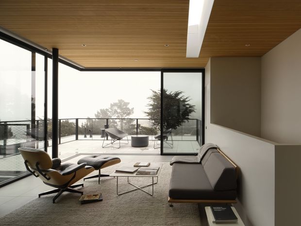 A refined contemporary living room with expansive black-framed glazing opening onto a terrace, a warm timber-lined ceiling, pale flooring, and a neutral seating arrangement. The room includes a sculptural lounge chair with ottoman, a streamlined sofa, a low glass coffee table, and a long recessed ceiling light. A prominent tree is visible beyond the terrace.