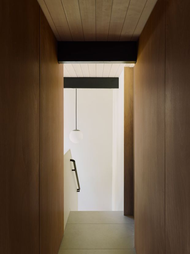 A narrow, minimalist interior passage framed by dark timber wall panelling and a matching timber-lined ceiling. The corridor opens toward a bright white stair or landing area, with a simple spherical pendant light and a partially visible black handrail. The composition emphasises strong vertical timber planes, concealed joinery, and contrasting light and dark surfaces.