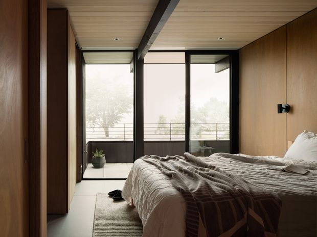 A warm, modern bedroom with a low-profile bed, neutral textured bedding, full-height glazing, and a balcony beyond. Dark timber cabinetry and wall panelling contrast with pale timber ceiling boards, while exposed dark structural beams frame the space. The room has a calm, refined atmosphere and strong indoor-outdoor connection.