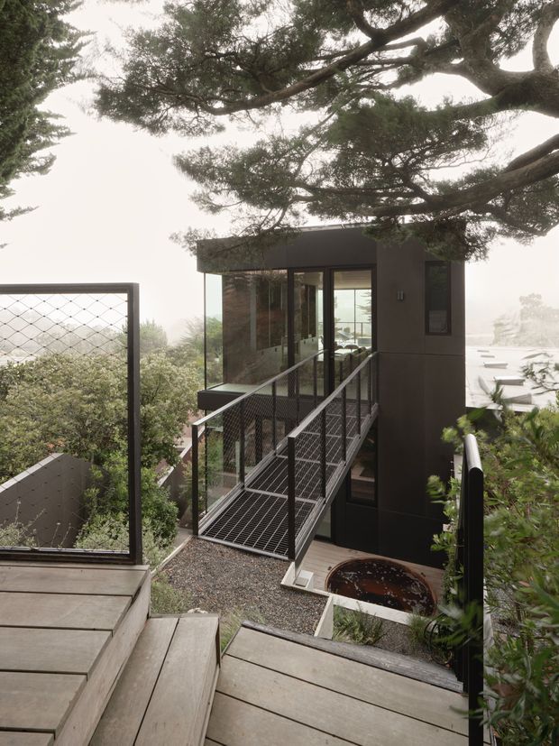 Exterior view of a compact modern pavilion or entry structure set into a lush, wooded sloping site. Dark metal cladding and extensive glazing form the small building, accessed by a steep grated metal bridge and framed by black mesh balustrades. Timber decking and steps occupy the foreground, with a partially visible circular spa or water feature below. Large mature tree branches canopy the scene.