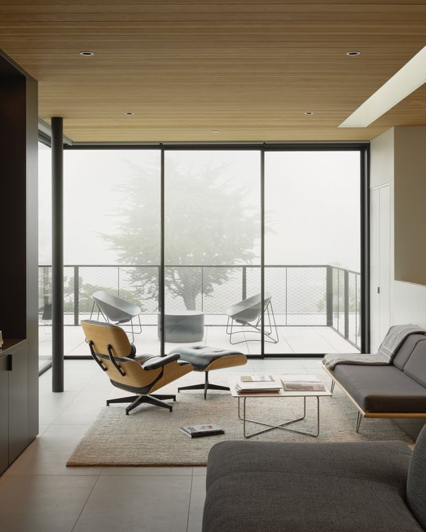 A refined contemporary living room with a low-profile grey sectional sofa, sculptural leather lounge chair and ottoman, and a slim metal-framed coffee table arranged on a textured neutral rug. Full-height black-framed glazing opens onto a balcony or terrace, while the warm timber-lined ceiling with recessed downlights and a long skylight adds visual warmth and natural light. The room has a calm, minimalist mid-century-influenced character.
