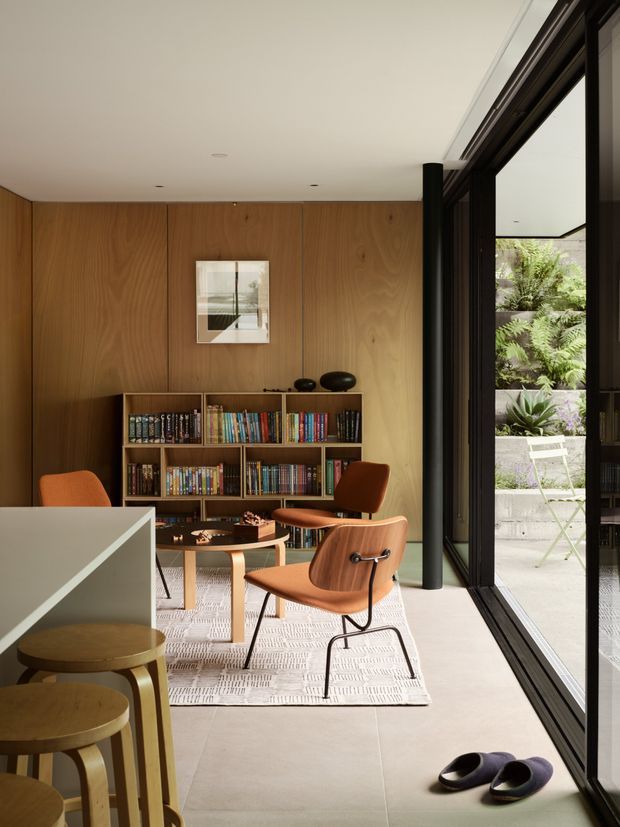 A warm, mid-century-inspired reading and living area with full-height timber wall panelling, a built-in open bookshelf filled with books, sculptural dark objects, and several orange-brown upholstered lounge chairs around a small round table. A large black-framed glazed opening connects the room to a planted outdoor courtyard. Pale large-format flooring, a light rug, and a simple white ceiling create a calm, refined interior.