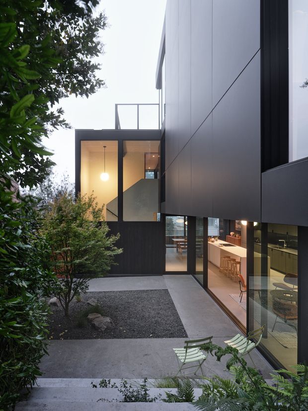 A contemporary multi-level home viewed from a landscaped courtyard. Dark cladding and large black-framed glazed openings define the facade, with a warmly lit open-plan kitchen and dining area visible through the ground-floor glazing. Concrete paths, a gravel garden bed, and dense planting surround the building. An upper-level terrace or balcony and an internal stair are also visible.
