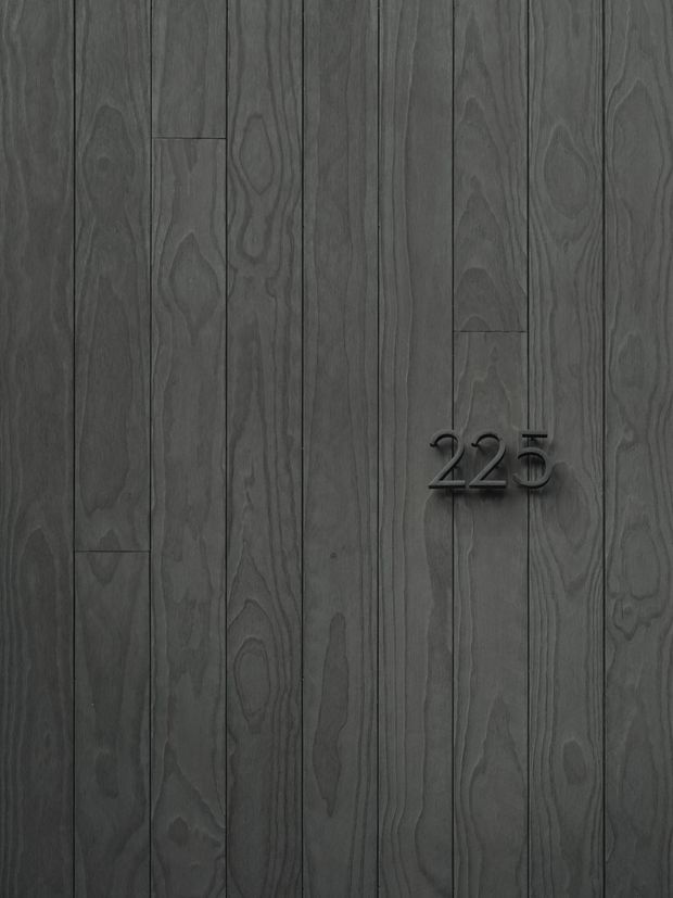 Close-up of a dark grey vertical timber wall cladding with visible natural grain and narrow board joints. Minimal black raised numerals reading “22B” are mounted near the center-right, suggesting a building entrance or address plaque.