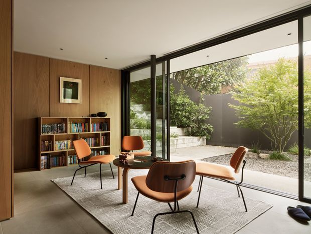 A refined mid-century-inspired sitting and reading area with a low wooden bookshelf against a full-height timber-paneled wall, several sculptural lounge chairs around a small round table, and a large patterned rug. Floor-to-ceiling black-framed glazing opens visually onto a minimalist enclosed courtyard garden with gravel, concrete planters, and trees. The space is softly lit, warm, and connected to the outdoors.