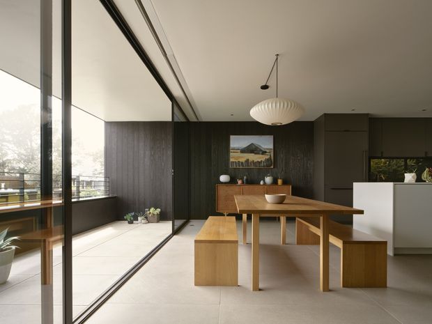 Contemporary open-plan dining area with a timber dining table, matching bench seating and stools, dark timber wall panelling, and a white kitchen island in the background. Full-height black-framed glazing opens onto a balcony or terrace, bringing in abundant natural light. A sculptural pendant light hangs above the table, with restrained decorative objects and indoor plants creating a calm, refined atmosphere.