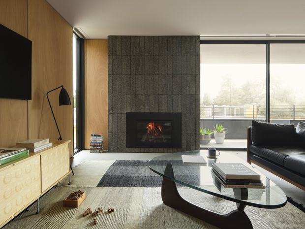 A refined contemporary living room with warm timber wall panelling, a textured dark stone fireplace surround, and large floor-to-ceiling glazing opening to a balcony. The room features a black leather sofa, sculptural glass coffee table, low timber media console, patterned rug, and understated mid-century-inspired furnishings. Natural daylight creates a balanced, inviting atmosphere.