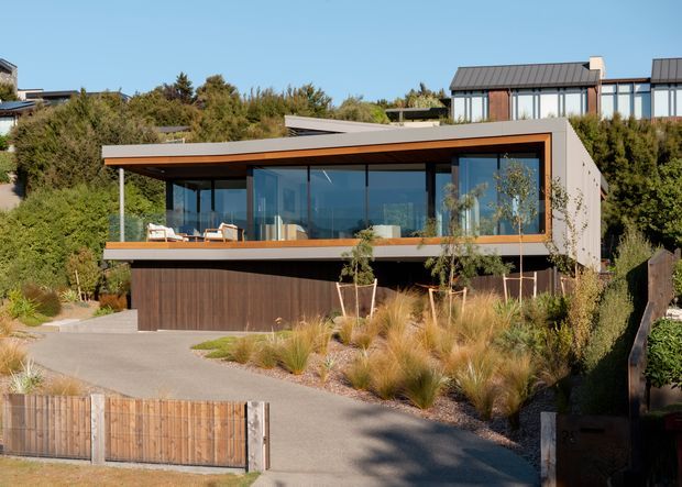 Exterior view of a contemporary, multi-level residential house on a landscaped sloping site. The home features a broad low-pitched roof, extensive floor-to-ceiling glazing, dark vertical timber cladding at the lower level, warm timber soffits and balcony trim, and a sheltered upper terrace. Native-style grasses, shrubs, timber fencing, and a driveway surround the building.