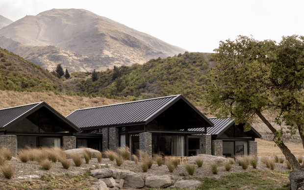 A low-profile contemporary mountain lodge set within a rugged alpine landscape. The building comprises several connected gabled volumes with dark ribbed metal roofs, extensive dark-framed glazing, and substantial rough stone-clad columns and walls. Native grasses, boulders, and scrub landscape surround the house, with a large bare mountain rising behind it.