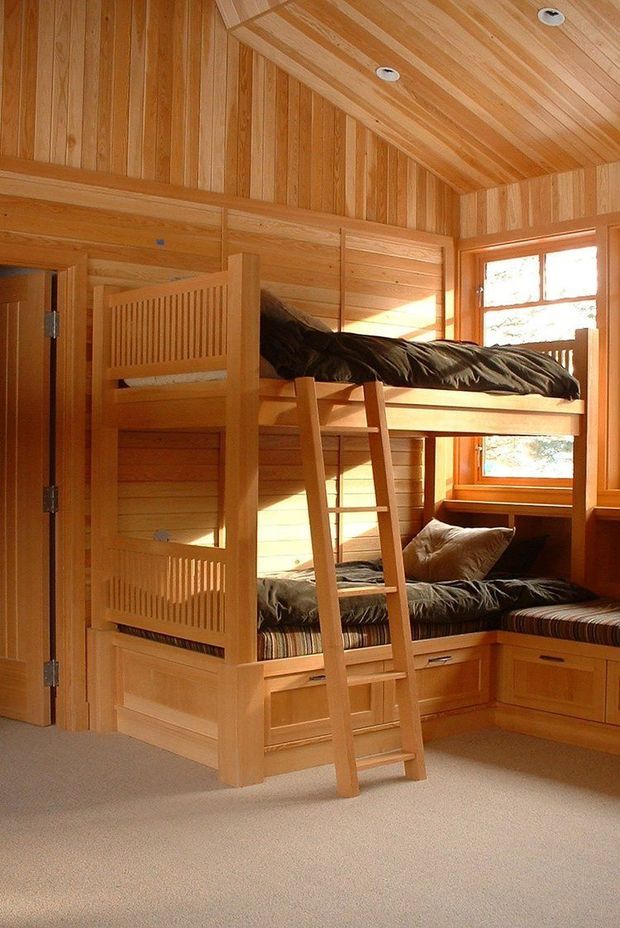 A warm, compact bunk bedroom lined extensively with honey-toned timber paneling on the walls and vaulted ceiling. Built-in wooden bunk beds feature integrated drawers and bench seating, with a central ladder, striped bedding, and a small window bringing in daylight. The room has soft beige carpet and a highly coordinated timber joinery scheme.