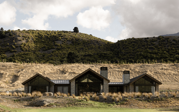 Single-storey rural lodge-style house set into a dry, rolling hillside. The low, symmetrical composition has several steep gabled volumes linked by dark metal-roofed sections, with dark-framed glazing, exposed stone walls, and a central recessed entry or living space. Native grasses and gravel landscaping surround the building, while scrub-covered hills and a partly cloudy sky form the background.