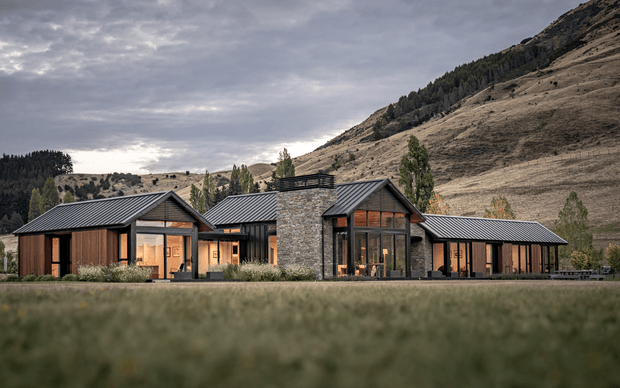 A low, modern mountain residence viewed across a blurred grassy foreground. The building is composed of several connected pitched-roof volumes with dark metal roofing, warm timber-clad walls, extensive dark-framed glazing, and a central stone-clad tower-like section. Steep, sparsely vegetated hills and woodland form the background under an overcast sky.