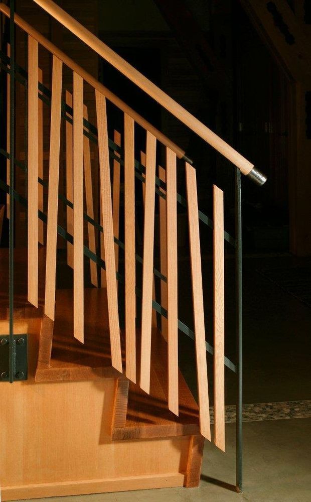 Close-up architectural photograph of a timber stairway with open vertical timber balusters, a diagonal timber handrail, dark metal supports, and timber-clad stair treads and risers. Dramatic warm sunlight highlights the wood against a dark interior background.