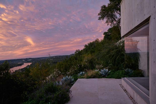 Dusk exterior view of a minimalist concrete residence set into a vegetated hillside, with a broad paved terrace, large horizontal glazing, and a distant waterway and settlement visible beyond the landscaped garden. The scene is dominated by dramatic pink and purple sunset light and a natural, secluded setting.