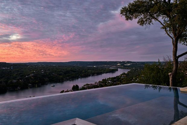 An elevated outdoor infinity pool overlooks a winding river and densely wooded residential landscape at dusk. The pool occupies the foreground, reflecting the pink and purple evening sky, while a mature tree frames the right side of the view.
