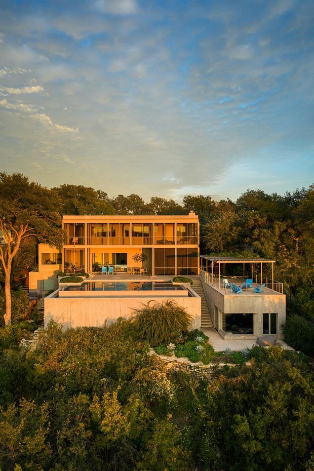 Exterior architectural photograph of a contemporary multi-level residence set within dense landscaped vegetation. The house has broad glazed openings, warm-toned rendered walls, flat rooflines, terraces and balconies, with a prominent rectangular swimming pool and a covered outdoor entertaining terrace. The image is captured in warm late-afternoon light, with the building integrated into a sloping garden site.