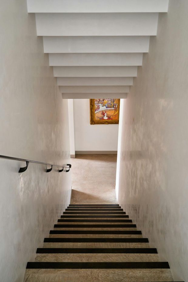 A narrow interior stairway descends toward a small landing. White textured walls frame dark stair treads, with a slim metal handrail and black brackets along the left wall. A series of white boxed ceiling beams creates a strong rhythmic pattern overhead, while a framed artwork is visible at the lower landing. The space is softly lit, neutral, and architecturally focused.
