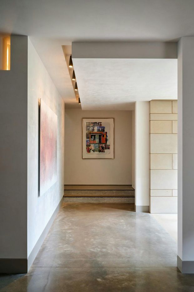 A minimalist interior entry hall or transition space with polished concrete flooring, warm neutral plaster walls, recessed linear lighting, and a short set of steps leading to a framed artwork. A built-in wall of pale rectangular panels is visible on the right, with a large abstract artwork on the left wall. The space has clean geometric volumes, soft indirect lighting, and a restrained material palette.