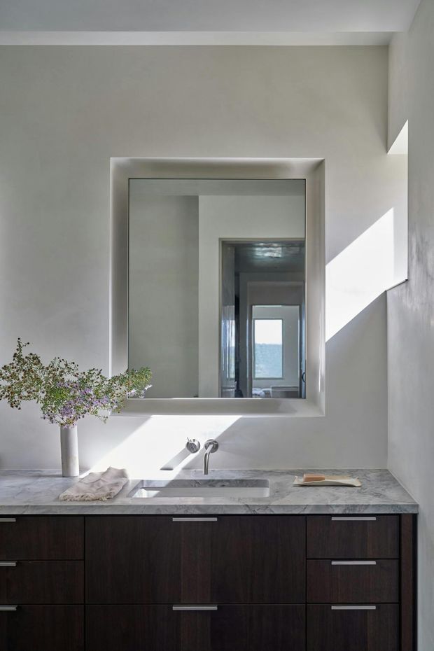 A refined bathroom vanity with a large recessed rectangular mirror, wall-mounted basin tapware, pale stone countertop, and dark timber cabinetry with integrated drawers. Strong sunlight creates geometric shadows across the light plaster walls and vanity. A small floral arrangement and folded towels add restrained styling. The mirror reflects a corridor or adjoining interior beyond.