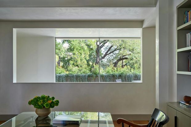 A refined, minimalist interior with a large horizontal window overlooking mature greenery. The room features smooth pale walls, a dark reflective table, a sculptural timber-and-black chair, and built-in shelving with books and a low cabinet. The composition is calm and light-filled, with a strong connection to the garden outside.
