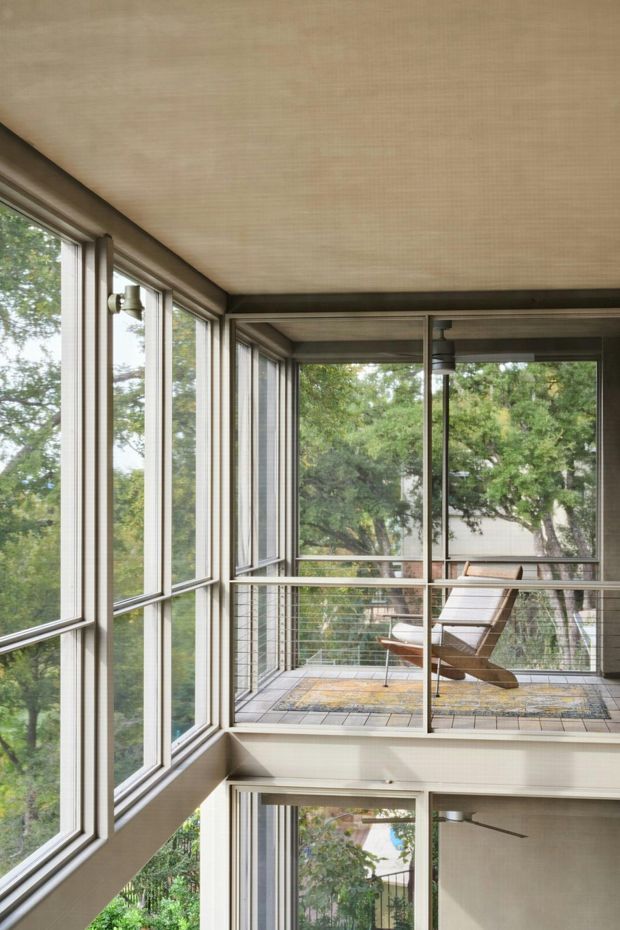 A bright enclosed glazed balcony or sunroom with extensive floor-to-ceiling windows, slim dark frames, and a light textured ceiling. A metal cable balustrade overlooks leafy trees, while a pair of mid-century-style lounge chairs and a patterned rug create a relaxed sitting area. The space appears connected to a multi-level interior with large windows and exposed structural framing.