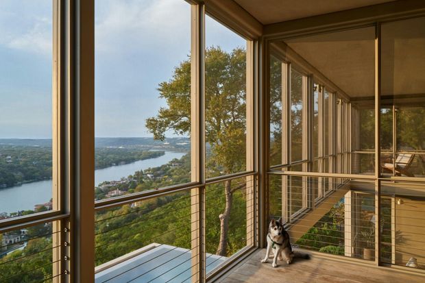 A covered elevated balcony or terrace enclosed with extensive floor-to-ceiling glazing and slim timber-framed windows, overlooking a winding river and leafy residential landscape. Warm timber decking, ceiling and framing combine with horizontal cable balustrades. A dog is present on the terrace, and the space extends along the building with a sheltered walkway-like form.