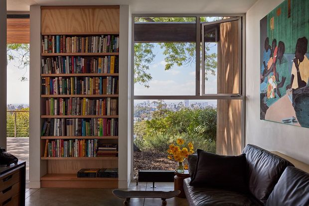 A compact, light-filled living room with a full-height built-in timber bookshelf, large operable windows, and a dark leather sofa. The windows overlook leafy vegetation and a distant urban landscape. A colourful artwork hangs on the pale wall, while a low coffee table and small side table complete the seating area.
