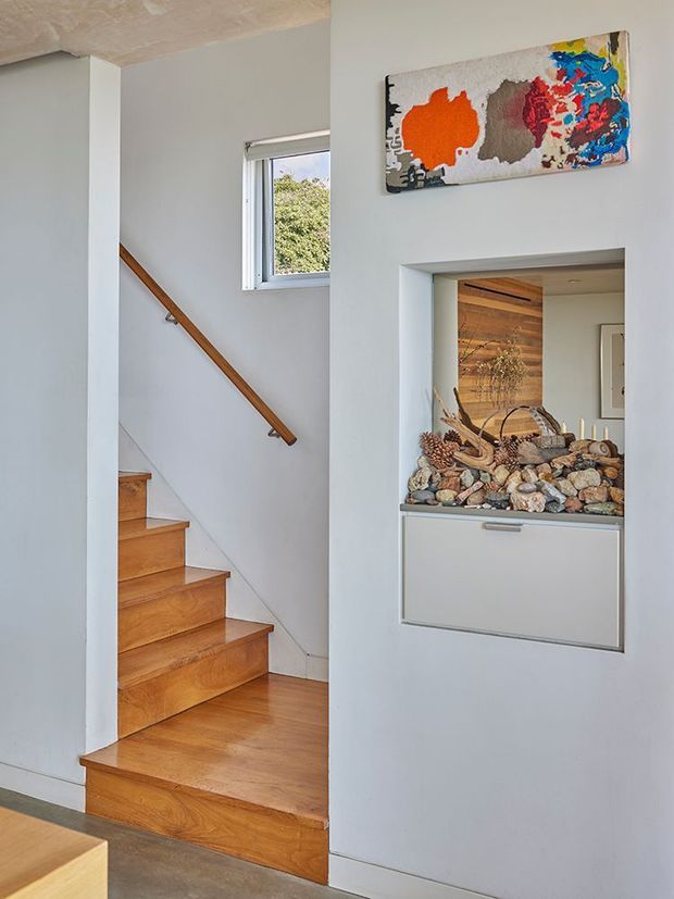 Interior view of a bright residential stairway with warm timber treads, flooring and handrail, white walls, and a small window. A recessed built-in display niche contains stones and driftwood, with a colourful abstract artwork mounted above. Another timber-lined room is visible through the doorway.