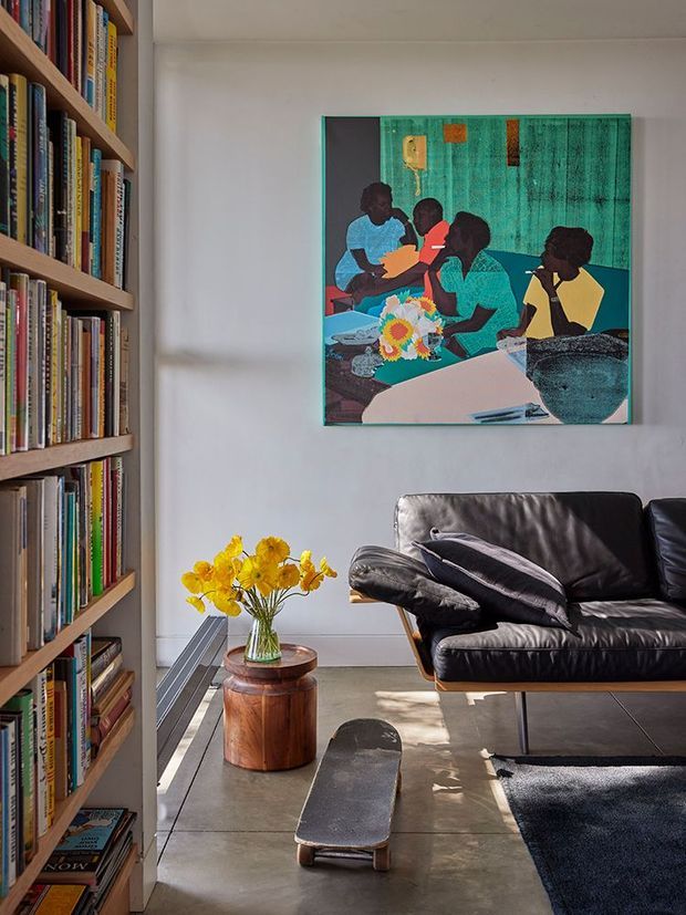 A bright, compact living room with a full-height built-in bookshelf packed with books along the left wall, a black leather sofa, a small round wooden side table with yellow flowers, a skateboard on the concrete-look floor, and a colourful contemporary artwork above the sofa. Natural light enters from the left, creating a relaxed, eclectic atmosphere.