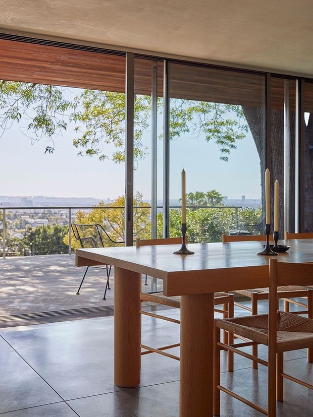 A refined dining area opens onto a broad terrace through floor-to-ceiling sliding glass doors. The room features a substantial warm timber dining table with cylindrical legs, woven timber dining chairs, dark metal candleholders, polished concrete flooring, and timber roller blinds. A distant urban landscape and trees are visible beyond the terrace, creating a bright indoor-outdoor connection.