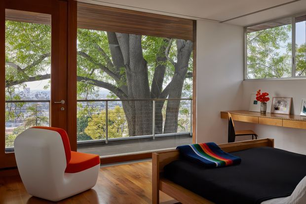 A bright, compact bedroom with a mid-century-inspired timber bed, black bedding, and a sculptural white-and-red lounge chair. Warm timber flooring and a full-height timber-framed glazed door and window open onto a balcony, with a large mature tree directly outside. A simple floating timber desk or shelf runs along the wall beneath the windows, displaying framed photographs and a small vase of flowers.