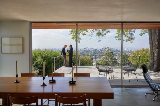 A refined mid-century-inspired dining area opens through expansive floor-to-ceiling glazing onto a spacious terrace with a city view. The interior features a long warm timber dining table, matching timber chairs, minimalist candleholders, smooth pale walls, and a low textured ceiling. Outside, a slim metal balustrade, tiled paving, and modern outdoor chairs sit beneath mature trees, creating a strong indoor–outdoor connection.
