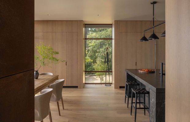 Warm, refined open-plan kitchen and dining space with full-height light timber cabinetry, wide timber flooring, a dark stone island with integrated seating, and black pendant lights. A narrow glazed opening provides a view to greenery beyond, while a timber dining table and upholstered chairs occupy the left side.