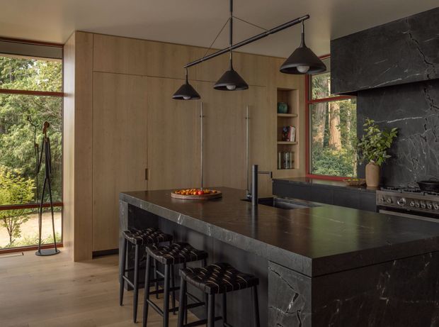 A refined contemporary kitchen with a large dark stone island, integrated seating for several people, timber cabinetry, black pendant lights, and a matching dark stone backsplash and range surround. Large timber-framed windows bring views of lush greenery into the space, while open shelving and understated styling add warmth.