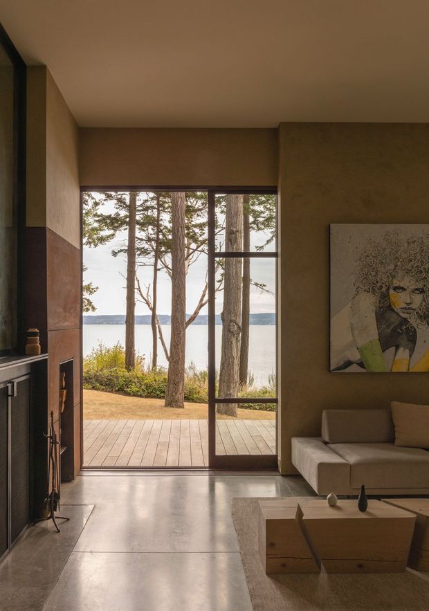 Warm, refined living room opening directly onto a timber deck and a wooded lakeside view. The room features a polished concrete floor, earthy plaster-like walls, a built-in fireplace with dark metal framing, large timber-framed glazing, a neutral sectional sofa, and a low timber coffee table. Natural light and muted brown tones create a calm, organic atmosphere.