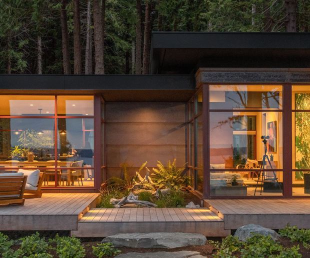 A contemporary single-storey forest retreat viewed from the exterior at dusk. Dark low-pitched roof planes frame extensive floor-to-ceiling glazing, with warm interior light revealing dining and living spaces. A timber deck wraps around a recessed planted courtyard containing ferns and rocks, with broad stone stepping elements leading through the landscaped foreground. The facade combines dark metal framing, warm timber or wood-toned cladding, and large glazed openings.