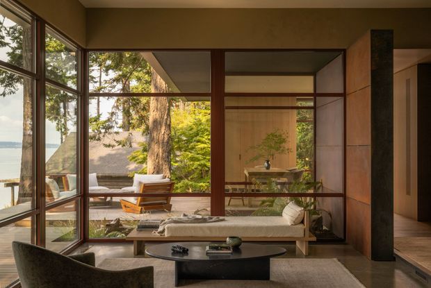 Warm, sophisticated living room with floor-to-ceiling timber-framed glazing and dark wood-framed internal glass partitions. The room includes a low built-in bench, sculptural dark coffee table, upholstered lounge chair, and a view through to a dining area and landscaped outdoor setting. Natural materials, strong horizontal lines, and abundant glazing create a calm connection between interior and exterior.
