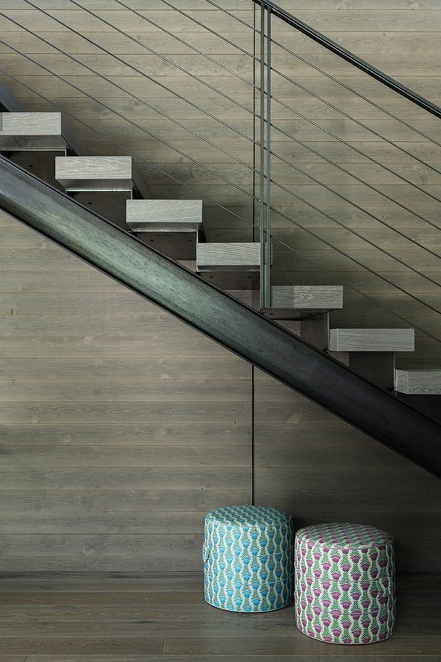 Close-up interior view of a modern floating staircase with open timber treads, a dark structural stringer, and thin horizontal metal cable balustrading. The staircase is set against matching horizontal timber wall panelling and flooring, with two colourful patterned cylindrical ottomans beneath it.